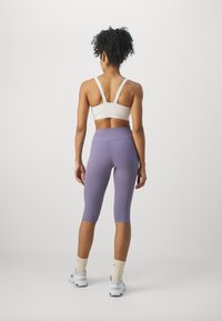 Bakifrån av en person som bär en beige sport-bh och lavendelfärgade högmidjade capri leggings, kompletterat med vita träningsskor och krämfärgade strumpor.