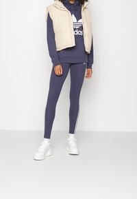 Marinblå sweatshirt med vit logotyp, beige vadderad väst, marinblå leggings med vita ränder och vita sneakers. Slät tyg, åtsittande design.