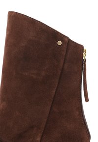 Gros plan sur une botte en daim marron avec une fermeture éclair dorée et un petit détail rond doré près du bord supérieur.