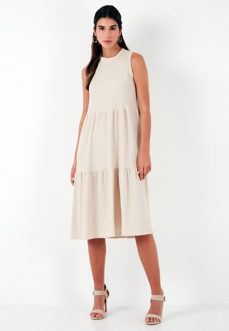 Beige sleeveless dress with a textured finish, featuring a round neckline and a tiered A-line silhouette. Paired with neutral high-heeled sandals.