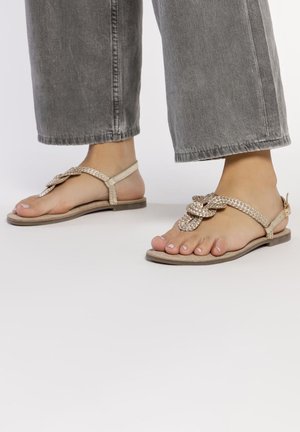 Sandalias planas beige con diseño de tira trenzada, que cuentan con un nudo decorativo y una pequeña tira trasera. Suela texturizada para un mejor agarre.