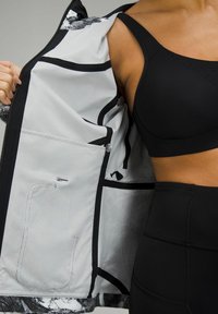 Personne portant un brassard de sport noir et un legging tient une veste ouverte montrant la doublure intérieure gris clair avec des détails de couture noirs.