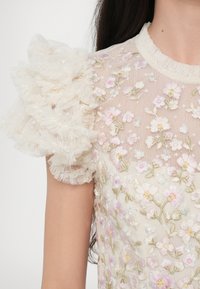 Top en dentelle crème transparente avec des fleurs roses et blanches brodées et une grande manche bouffante en dentelle à volants sur l'épaule droite.