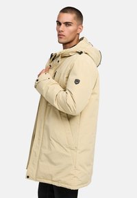 Beige vadderad parka med huva, utrustad med dragkedja och knappstängningar, justerbara snören och en logotypapplikation på vänster ärm. Slät textur.