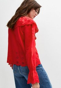 Blouse en coton rouge avec broderie à l'œillet, col à festons et longues manches avec des poignets volantés, associée à un jean en denim bleu.
