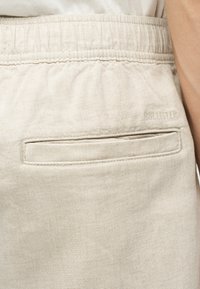 Μπεζ υφασμάτινα σορτς με ελαστική μέση και μία πίσω τσέπη τύπου welt, φορεμένα από άτομο με ορατό χέρι.