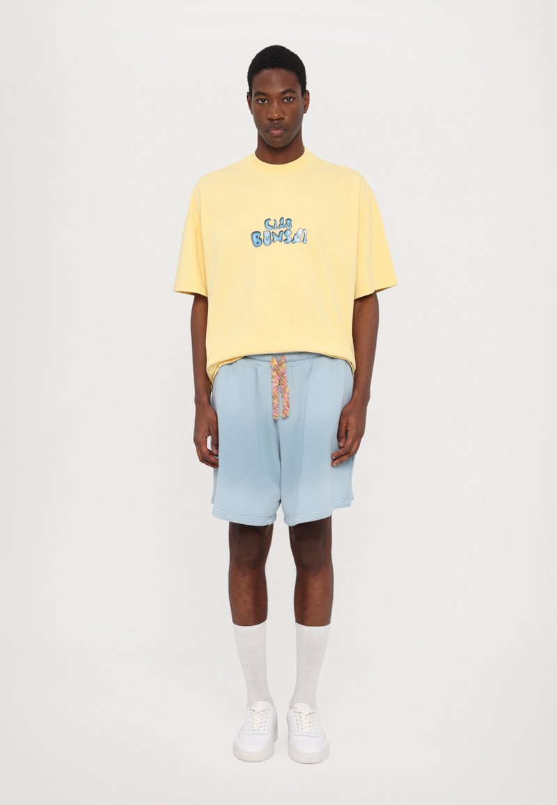 Geel t-shirt met korte mouwen en blauwe graphic tekst "Ciao Bonsai." Lichtblauwe shorts met een bloemenkoord. Witte sneakers en grijze sokken.