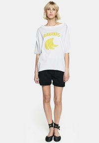 Biały, oversize'owy t-shirt z żółtym nadrukiem i grafiką "BANANAS", zestawiony z czarnymi szortami. Płaskie czarne buty z wiązaniami na kostce.