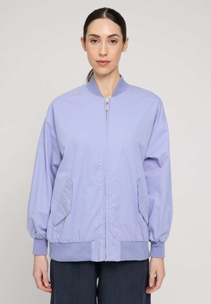 Woman in a light purple zip-up bomber jacket with pockets, standing against a plain light background, facing forward.