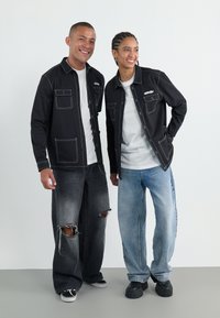 Schwarze Jacken mit weißem Stich, zwei Brusttaschen, graue zerrissene Jeans und blaue weit geschnittene Jeans. Beide tragen weiße T-Shirts und schwarze Schuhe.