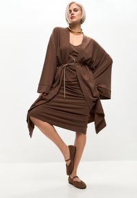 Vestido de tela marrón con un diseño envolvente, superpuesto con un cárdigan holgado y oversized. Incluye una cintura anudada y zapatos planos marrones.