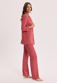 Kobieta stojąca bokiem, ubrana w koralowo-różową, dopasowaną marynarkę i pasujące szerokie spodnie oraz beżowe szpilki z ostrym noskiem.
