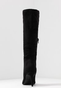 Bota de tacón stiletto de ante negro hasta la rodilla con cremallera lateral, vista desde la parte trasera sobre una superficie blanca y fondo liso.