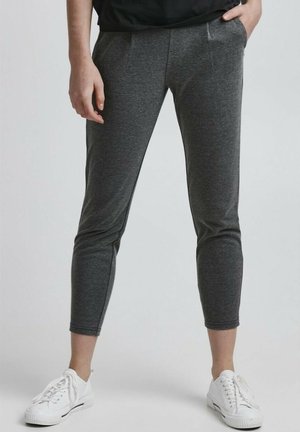 Person trägt dunkelgraue, enganliegende, knöchellange Hosen mit Seitentaschen und weiße niedrige Sneaker, steht vor einfachem Hintergrund.