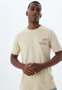 Beige katoenen t-shirt met korte mouwen, ronde halslijn en geborduurde tekst "LIFE" en "SOUL" in bruin. Soepele textuur, casual pasvorm.