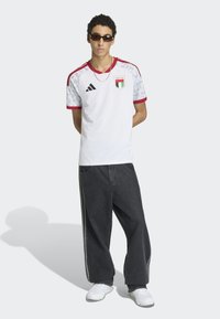 Maillot de football blanc avec des accents rouges et gris, présentant un patch et le logo Adidas, associé à un jean noir ample et des baskets blanches.