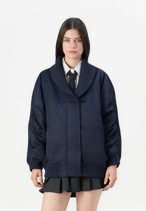 Femme aux cheveux noirs lisses portant une veste oversize bleu marine, une chemise rayée, une cravate noire et une jupe plissée foncée sur fond blanc.