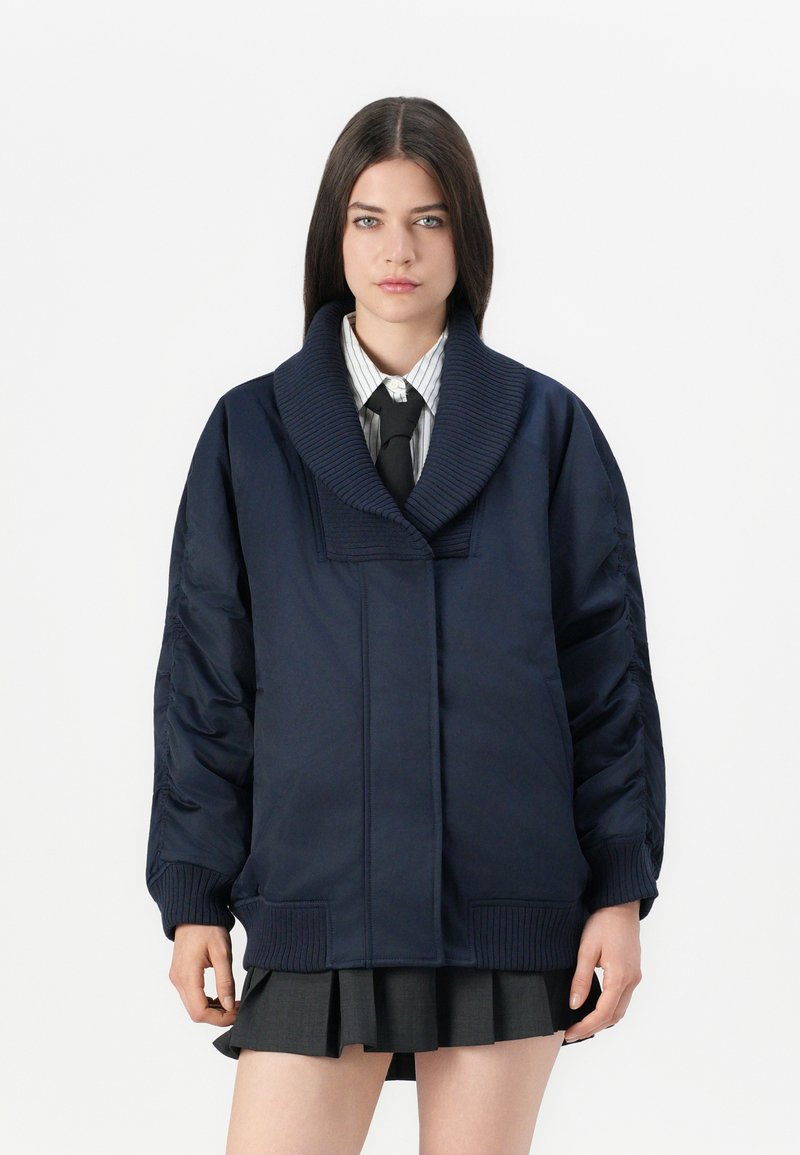 Donna con capelli scuri e lisci che indossa un giubbotto oversized blu navy, una camicia a righe, una cravatta nera e una gonna plissettata scura su uno sfondo bianco.