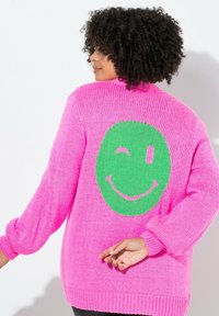 Maglione lavorato a maglia spesso di un brillante colore rosa, con maniche lunghe. Presenta un grande motivo di faccina sorridente verde sul retro. Fit strutturato e rilassato.