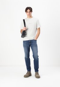 T-shirt grigio a maniche corte con logo sottile, jeans blu e stivali beige con lacci. Borsa a tracolla nera indossata attraverso il corpo.
