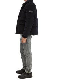 Giubbotto imbottito blu con colletto alto, tessuto strutturato e tasche laterali. Abbinato a jeans grigio chiaro e scarpe slip-on nere con un accento verde.