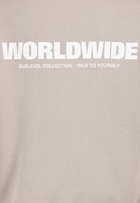 Beige sweatshirt met gedurfde witte tekst: "WORLDWIDE SUBLEVEL COLLECTION - TRUE TO YOURSELF." Zacht, gestructureerd materiaal met een losse pasvorm.
