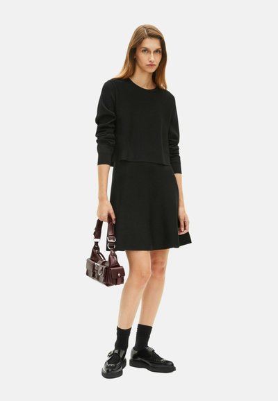 Zwarte cropped sweatshirt gecombineerd met een mouwloze zwarte jurk. Heeft een glanzende bordeauxrode tas en zwarte schoenen met dikke zolen en sokken.