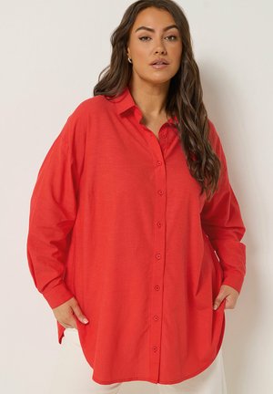 Kvinde med langt mørkt hår iført en løs rød skjorte med knapper og hvide bukser, stående med hænderne i lommerne foran en ensfarvet baggrund.