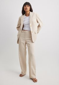 Beige Blazer mit zwei Knöpfen und einer Brusttasche, kombiniert mit passenden hoch-taillierten Hosen, gestylt mit einem weißen T-Shirt und Sandalen.