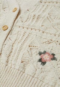 Kremowy sweter z dzianiny, z teksturowanym wzorem, drewnianymi guzikami oraz wyszywanymi w kwiaty detalami w kolorze różowym i zielonym.