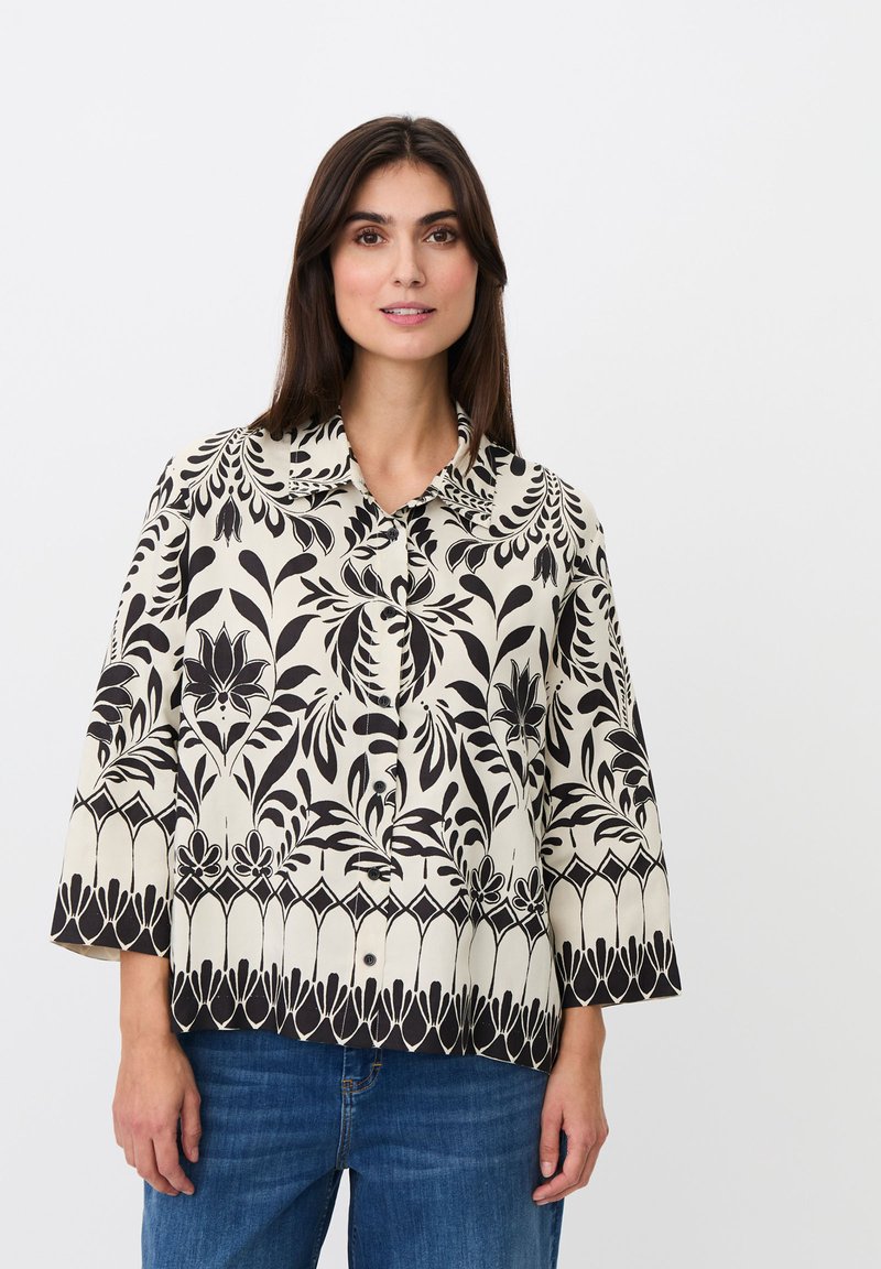 Femme portant un chemisier crème boutonné avec des motifs floraux noirs et des manches larges, associé à un jean bleu, debout devant un fond uni.