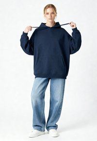 Sweat à capuche oversize bleu marine avec une poche avant, une capuche à cordon de serrage et des poignets côtelés, associé à un jean pattes d'éléphant bleu clair et des baskets blanches.