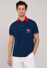 Marineblaues Poloshirt mit rotem Kragen, ausgestattet mit einer Brusttasche und gestickten Details. Kurze Ärmel mit weißen und roten Akzenten an den Bündchen.