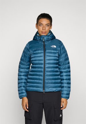 Blauwe pufferjack met verticale gestikte naden, een capuchon en een rits aan de voorkant. Heeft een logo op de linkerkant van de borst en een gladde textuur.