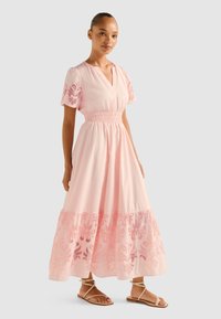 Rochie maxi din bumbac roz, cu mâneci scurte, talie elastică, decolteu în V și detalii florale brodate la tiv. Poartă sandale ușoare.