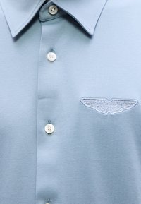 Ανοιχτό μπλε κοντομάνικο πουκάμισο με μυτερό γιακά, λευκά κουμπιά και κεντημένο λογότυπο Aston Martin στο αριστερό στήθος.