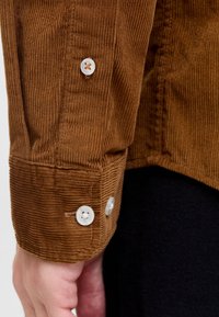 Brun manchester-skjortärm med strukturerade räfflor, två vita knappar och en skräddarsydd passform, mot en svart tygbakgrund.