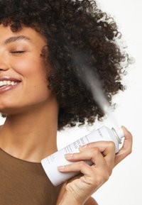 Canette de spray capillaire aérosol blanche tenue dans une main, avec un spray de couleur grise. Des cheveux bouclés et un haut sans manches de teinte neutre sont visibles en arrière-plan.