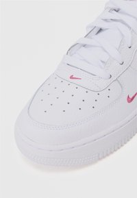 Valkoinen nahkakenkä, jossa on rei'itetty varvasosa, tasaiset nauhat ja vaaleanpunainen brodeerattu Nike-logokuva sivulla. Teksturoitu pohja, jossa on liukuestokuosi.