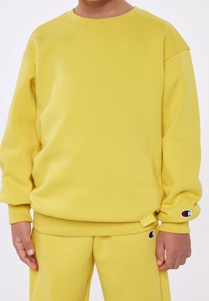 Barn iført matchende gul sweatshirt og bukser med Champion-mærkets logoer på ærmet og buksebenet.
