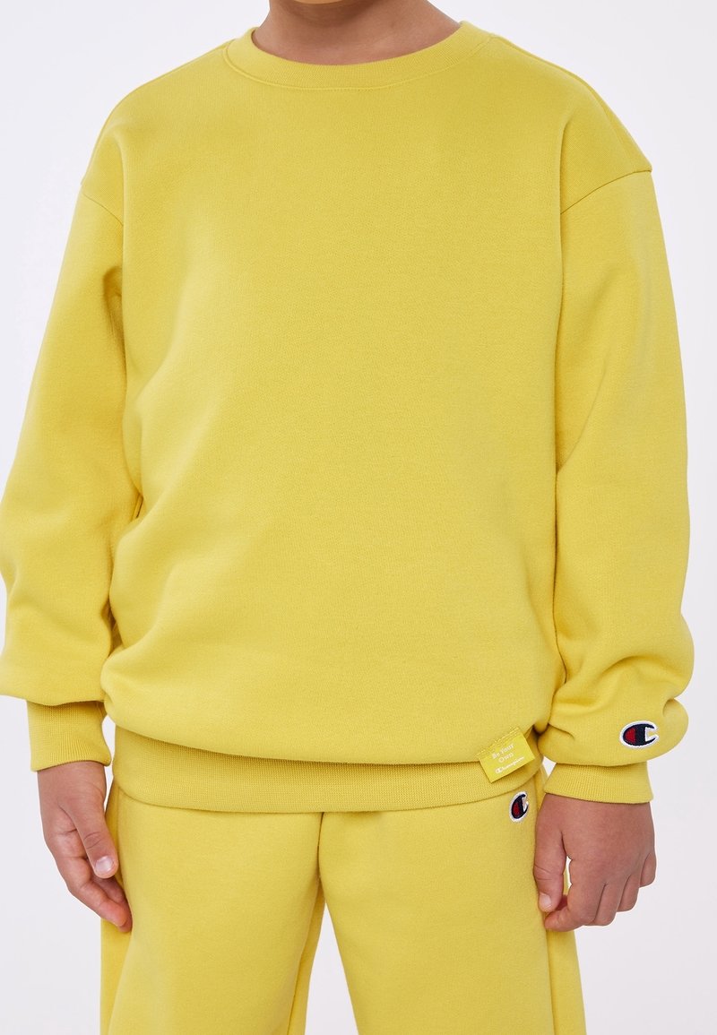 Enfant portant un sweat-shirt jaune assorti et un pantalon avec des logos de la marque Champion sur la manche et la jambe du pantalon.