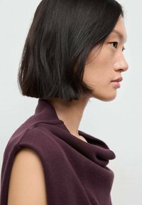 Giovane donna con i capelli neri corti di profilo che indossa un top senza maniche di un viola scuro su uno sfondo chiaro e semplice.