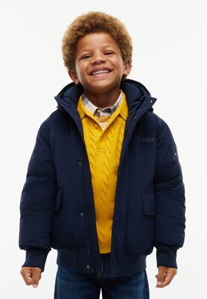 Lächelnder Junge mit lockigem Haar, der eine marineblaue Jacke, einen gelben Strickpullover und ein kariertes Hemd vor weißem Hintergrund trägt.