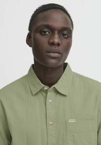 Camicia verde chiaro con bottone, realizzata in tessuto testurizzato, colletto a punta e taschino sul petto. Presenta bottoni bianchi e un piccolo etichetta con logo.