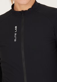 Musta urheilutakki, jossa on korkea kaulus, edessä vetoketju ja slim fit -design. Vasemmalla puolella on valkoinen "ELITE LAB" -logo.