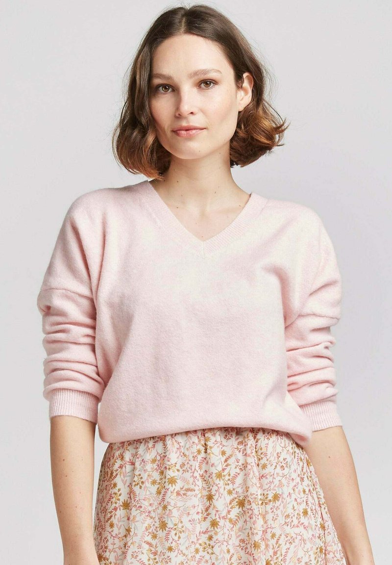 Pull à col en V rose clair en tissu doux et texturé avec des manches lâches, assorti à une jupe à motifs floraux aux couleurs pastel.