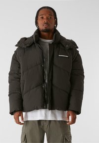Schwarze Puffjacke mit hohem Kragen, diagonalen Nähten und einem Frontreißverschluss. Verfügt über ein Logo-Patch im linken Brustbereich.