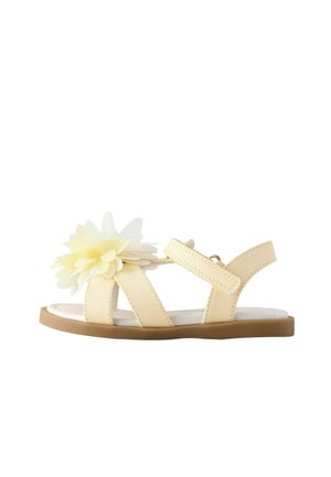 Béžová detská sandála s nastaviteľným remienkom a veľkým ozdobným bledozeleným látkovým kvetom na vrchu, plochá hnedá podrážka.