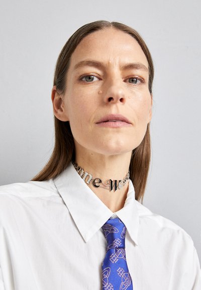 Valkoinen kauluspaita päällä käytettävä hopeinen kaulapanta, jossa lukee "some" ja "one". Sinisessä solmiossa on hienovarainen geometrinen kuvio.
