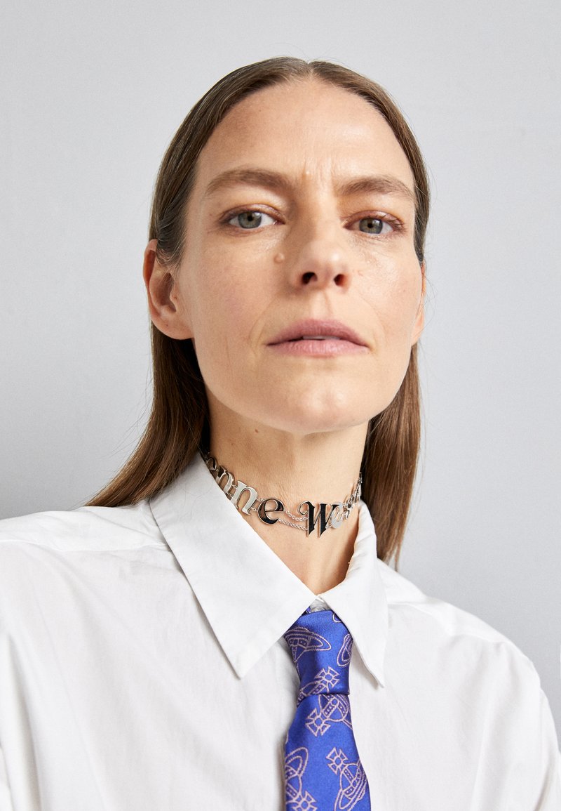 Silver choker with letters "some" and "one," worn over a white collared shirt. A blue tie features a subtle pattern with geometric shapes.